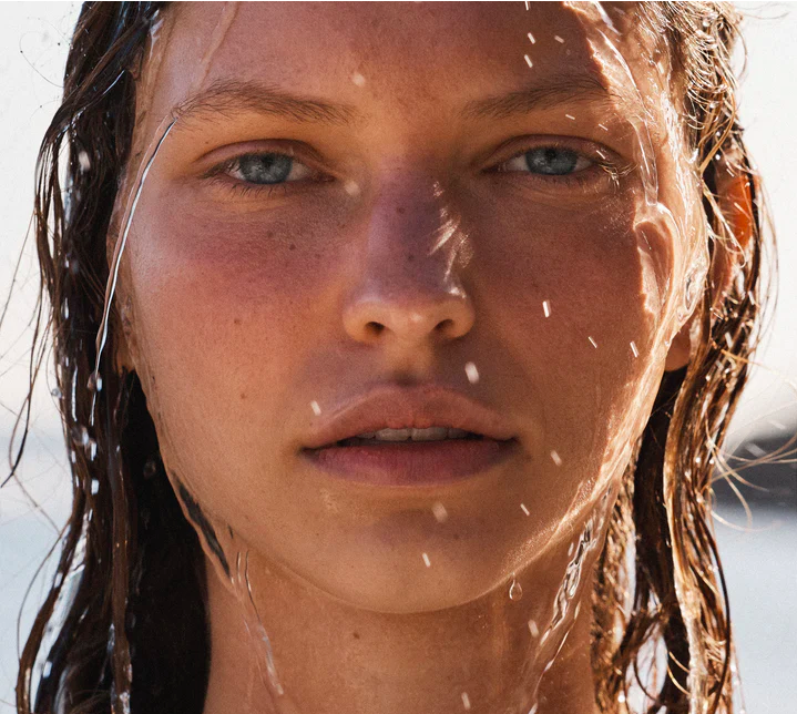Close-up portrait of a fair-skinned adult woman with blue eyes, wet hair, and water droplets on her face, bathed in natural light.