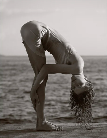 Monochrome image of a woman bending forward, head near the ground, standing on a stone surface with a blurred ocean background.