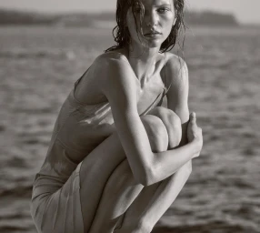 Model with wet hair in a slip dress crouching by the ocean for Beauty Department Under The Sun project.