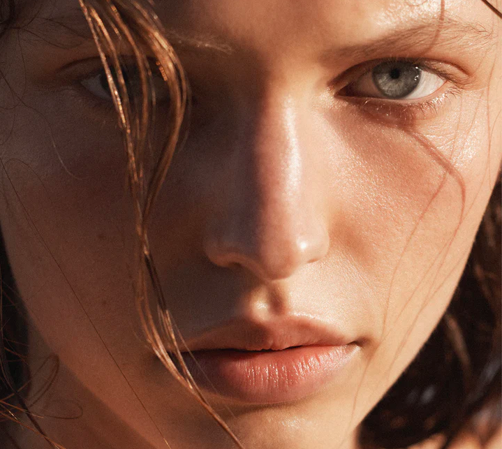 Close-up portrait of a young fair-skinned adult woman with wet brown hair, subtle makeup, and grey eyes in warm, natural light.