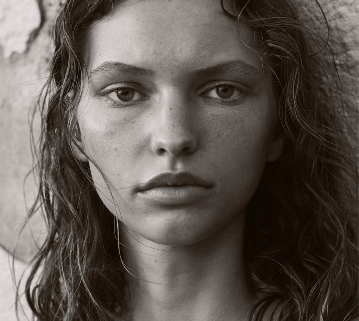 Close-up portrait of a young adult woman with wet, wavy hair, gazing directly at the camera against a textured wall backdrop.