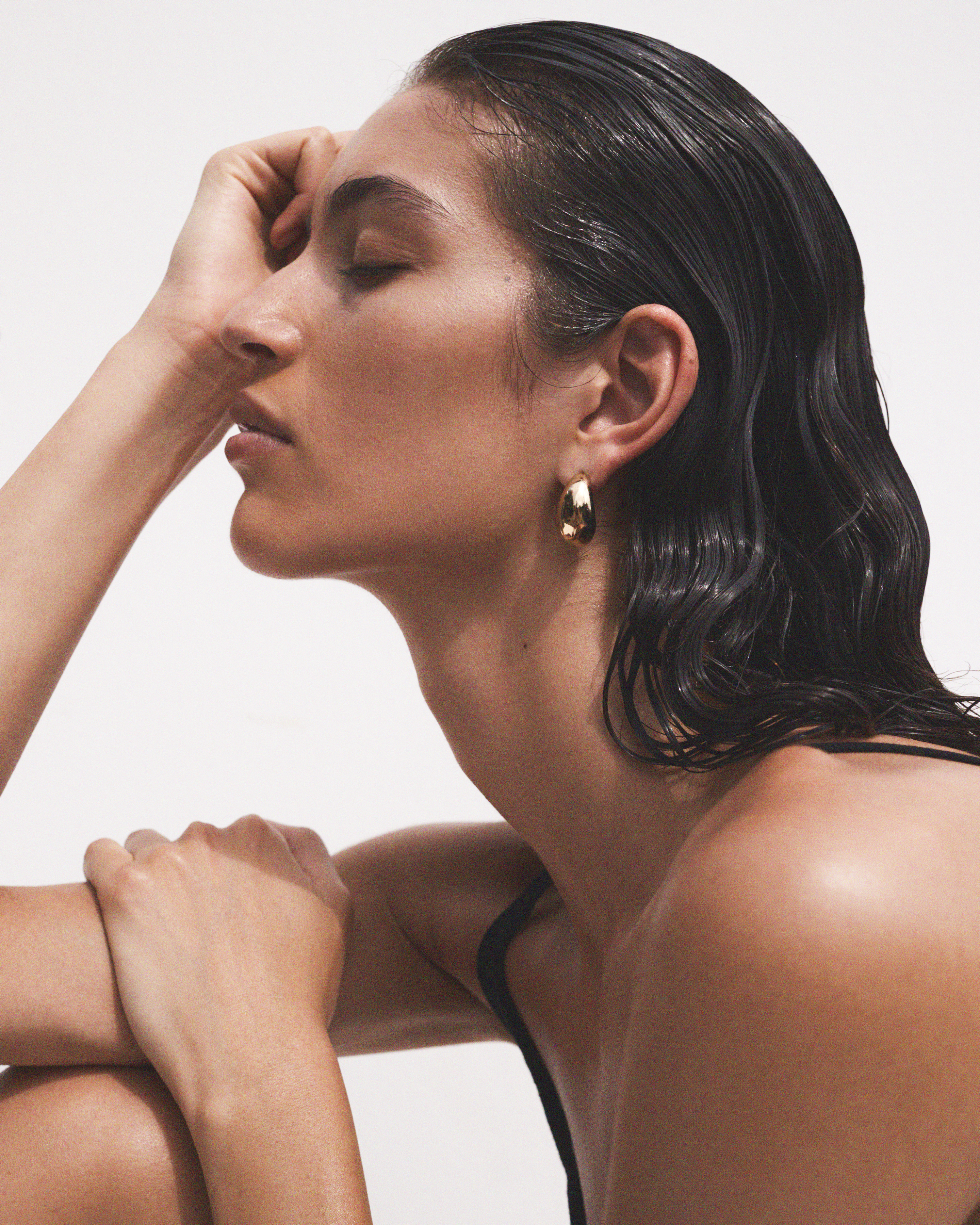 Profile portrait of a woman with her eyes closed and hand on her head, featuring gold hoop earrings, glossy hair, and a bright white background.
