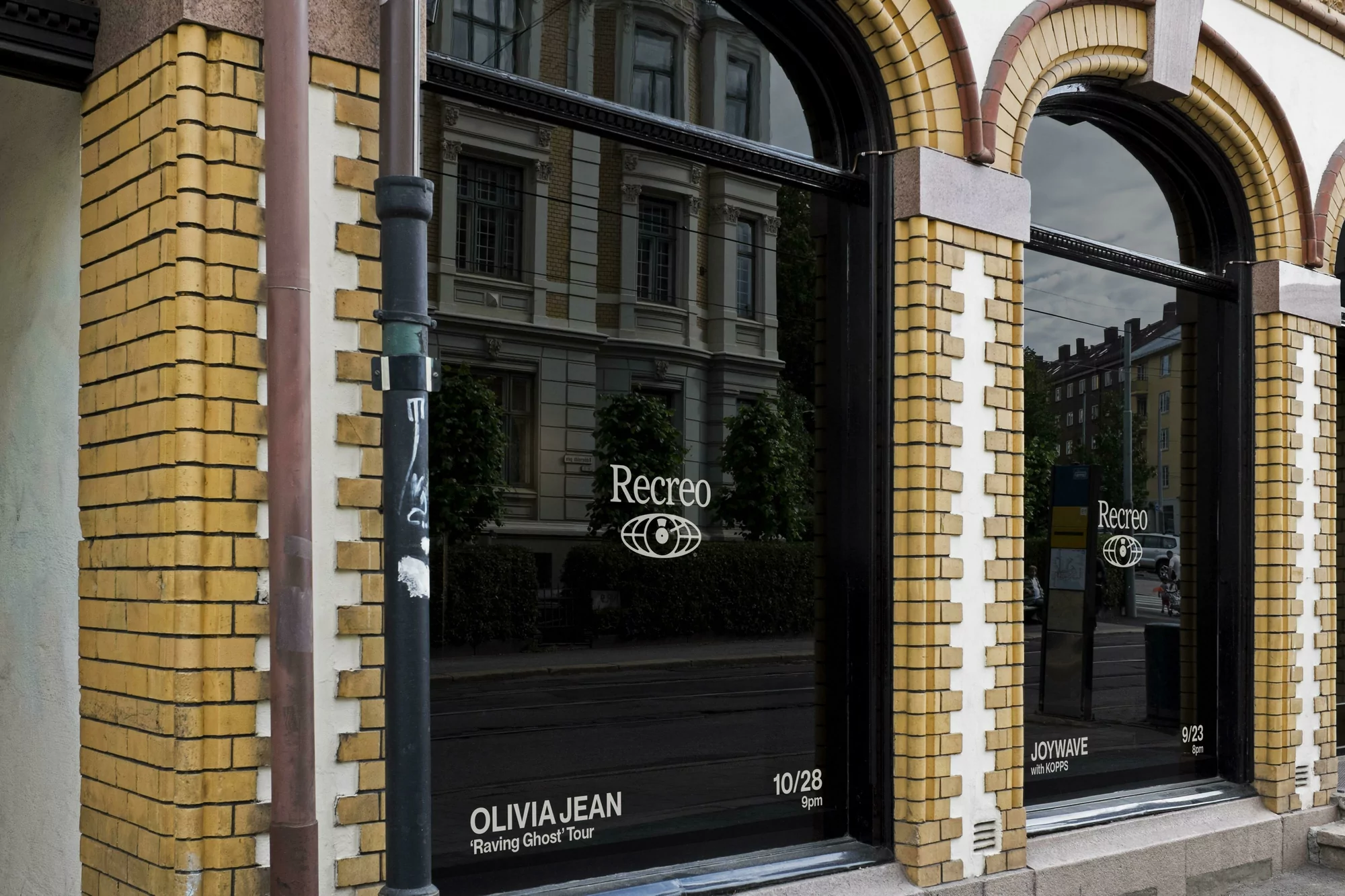 Exterior view of a building with arched windows displaying posters for upcoming events; street pole on the left.