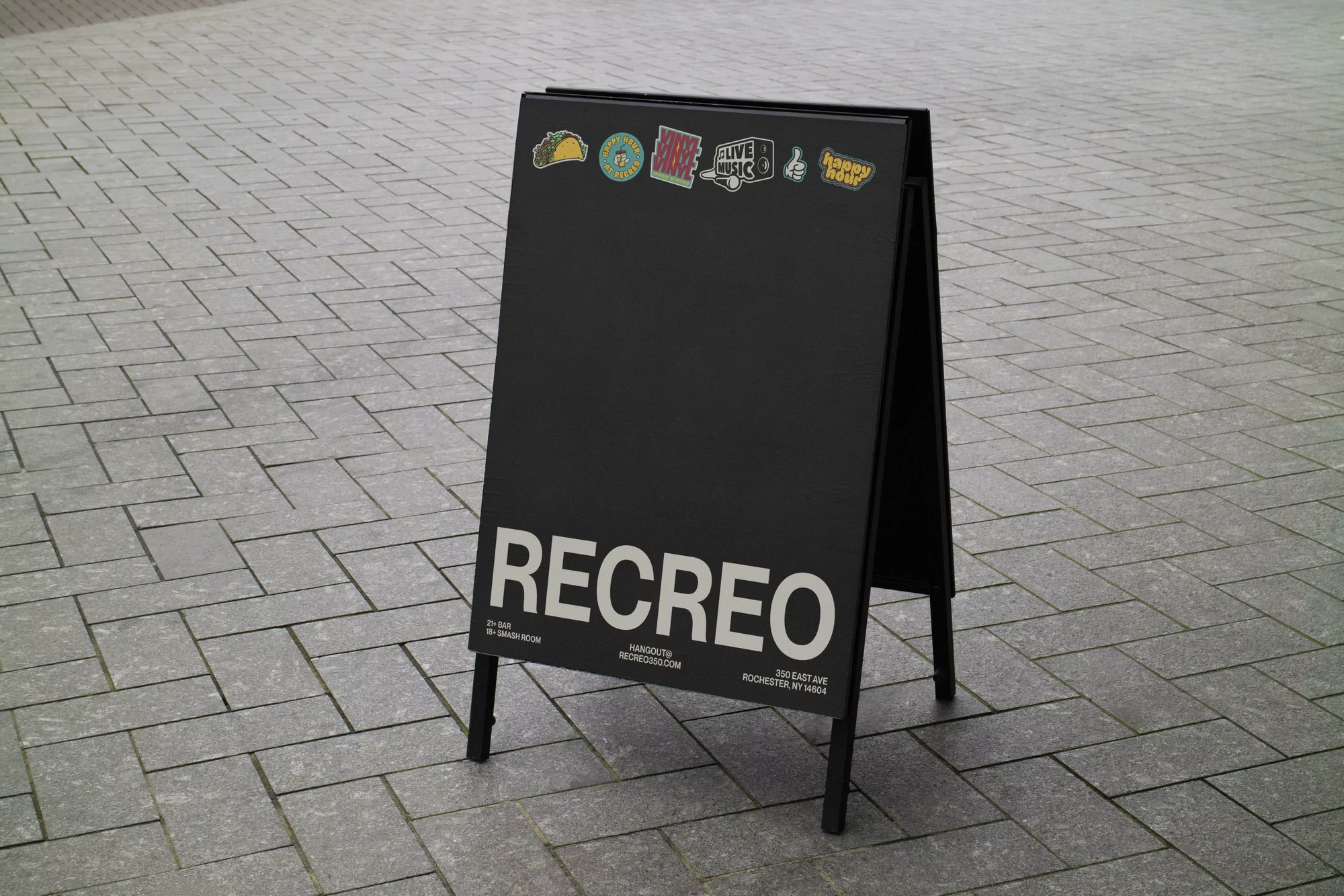 Black A-frame sidewalk sign featuring the brand name RECREO in white block letters, displayed on grey paving stones.