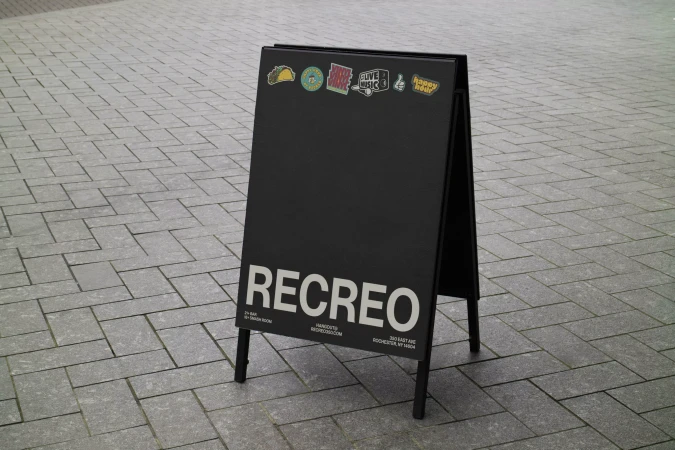 Black A-frame sidewalk sign featuring the brand name RECREO in white block letters, displayed on grey paving stones.