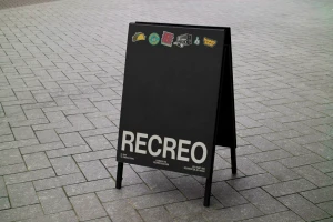 Black A-frame sidewalk sign featuring the brand name RECREO in white block letters, displayed on grey paving stones.