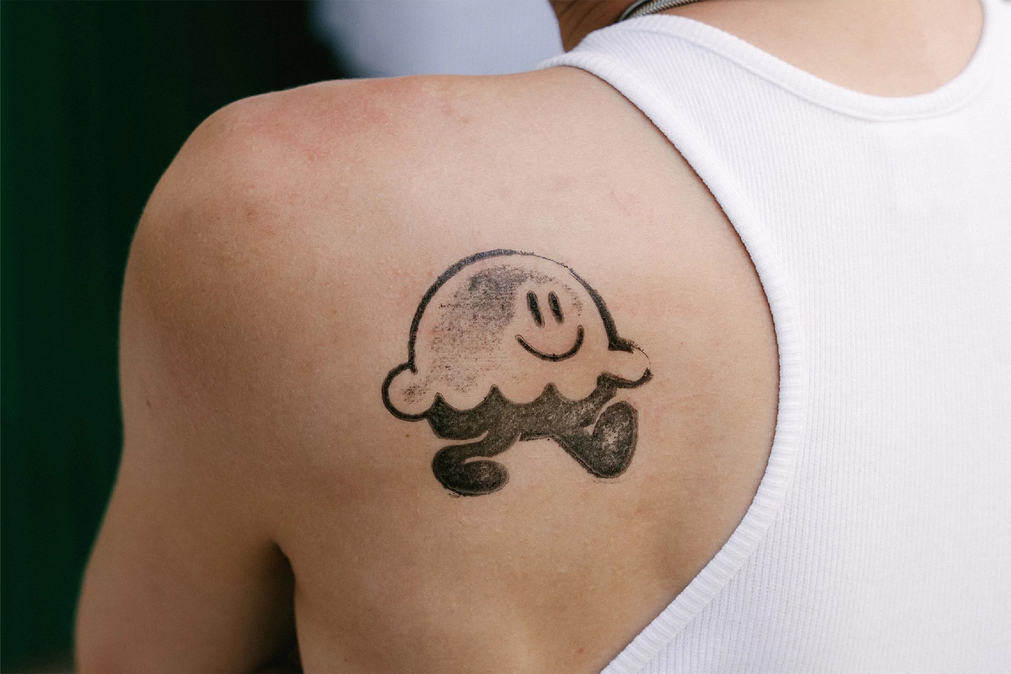 Close-up of a person's back and shoulder featuring a tattoo of a black cartoon character with a smiling face and small feet.