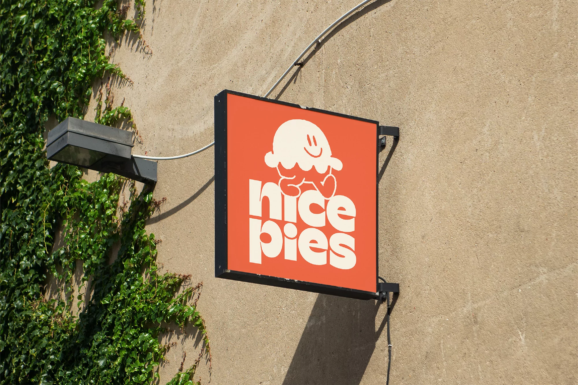 Orange sign that reads 'Nice Pies' with a cartoon pie character, mounted on a textured wall next to vibrant green ivy.