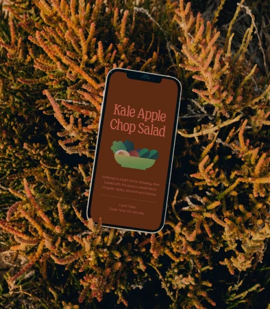 Smartphone displaying a recipe for Kale Apple Chop Salad, resting on a bed of orange succulent plants, creating a natural and inviting food image.