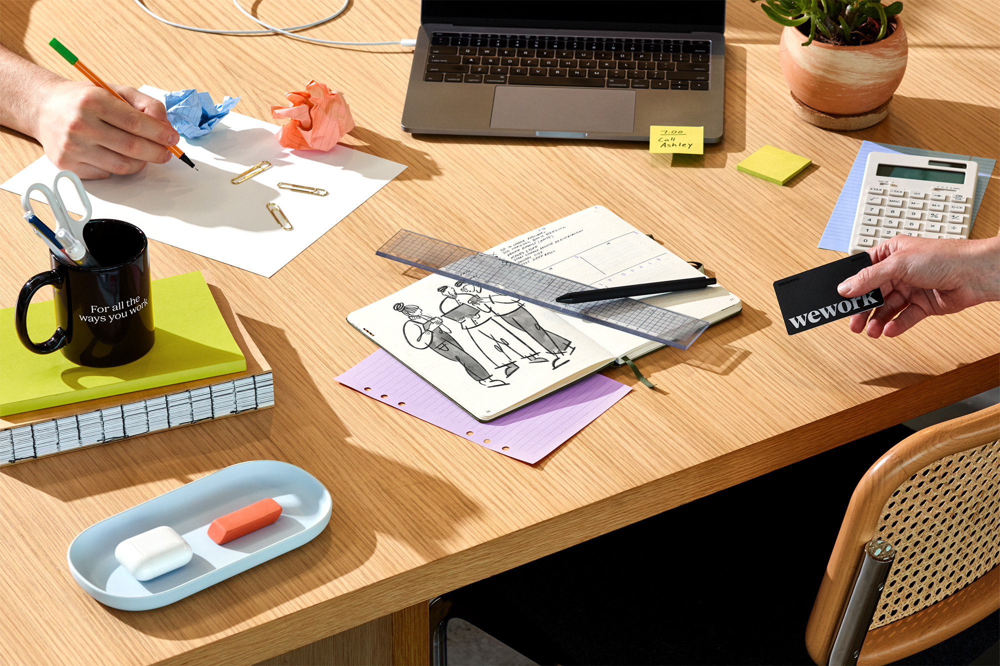 Overhead shot of a light wood desk with a laptop, notebooks, pens, and office supplies, and hands holding a black WeWork card.