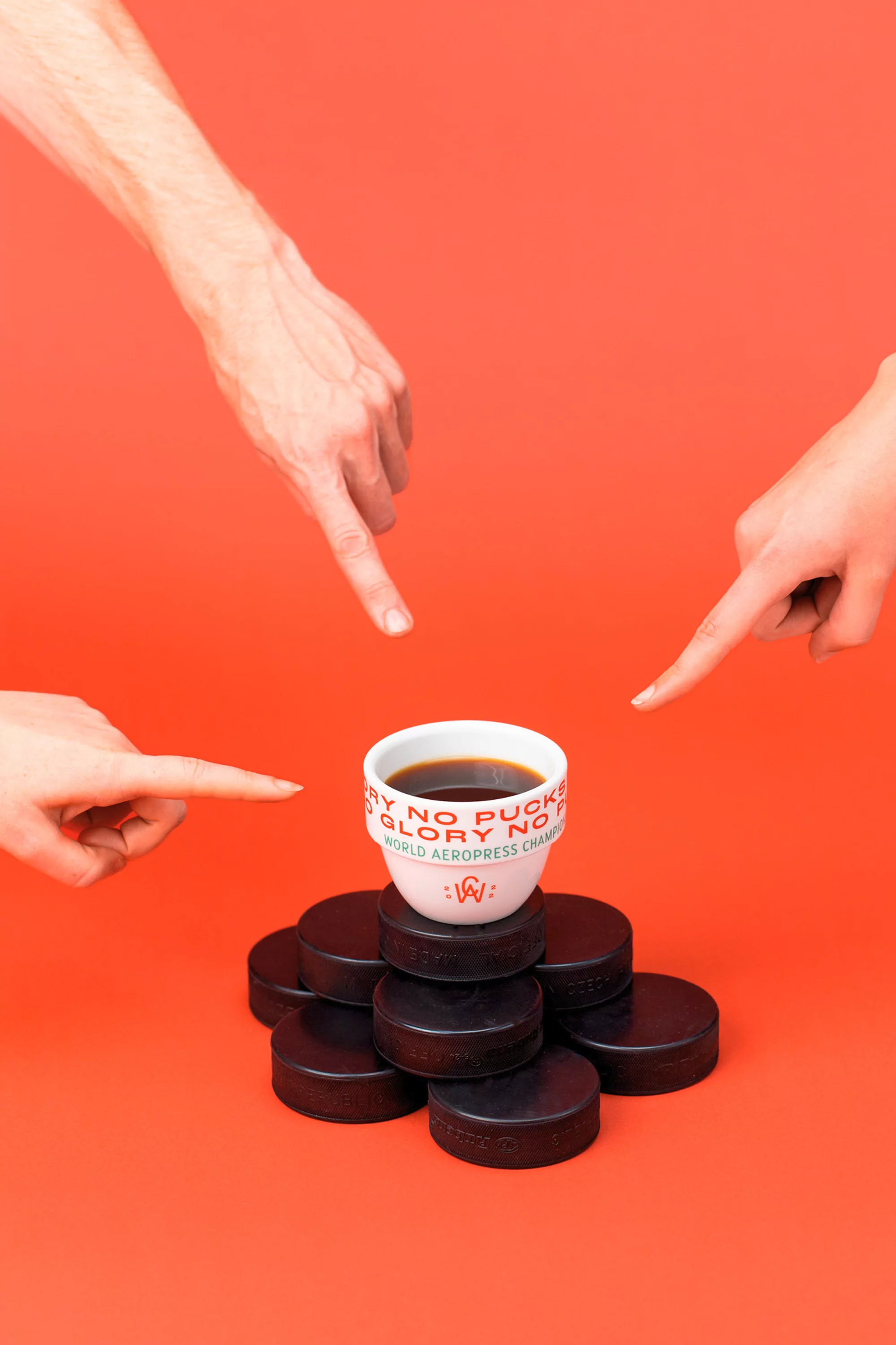 Studio shot of a cup of coffee on hockey pucks, pointed to by three hands, against an orange background, studio lighting.