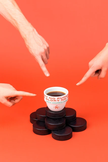 Studio shot of a cup of coffee on hockey pucks, pointed to by three hands, against an orange background, studio lighting.