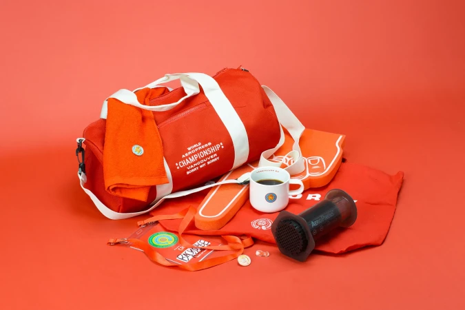 Promotional image features Aeropress coffee maker, mug, orange duffel bag, and fan-shaped foam hand on a coral background.
