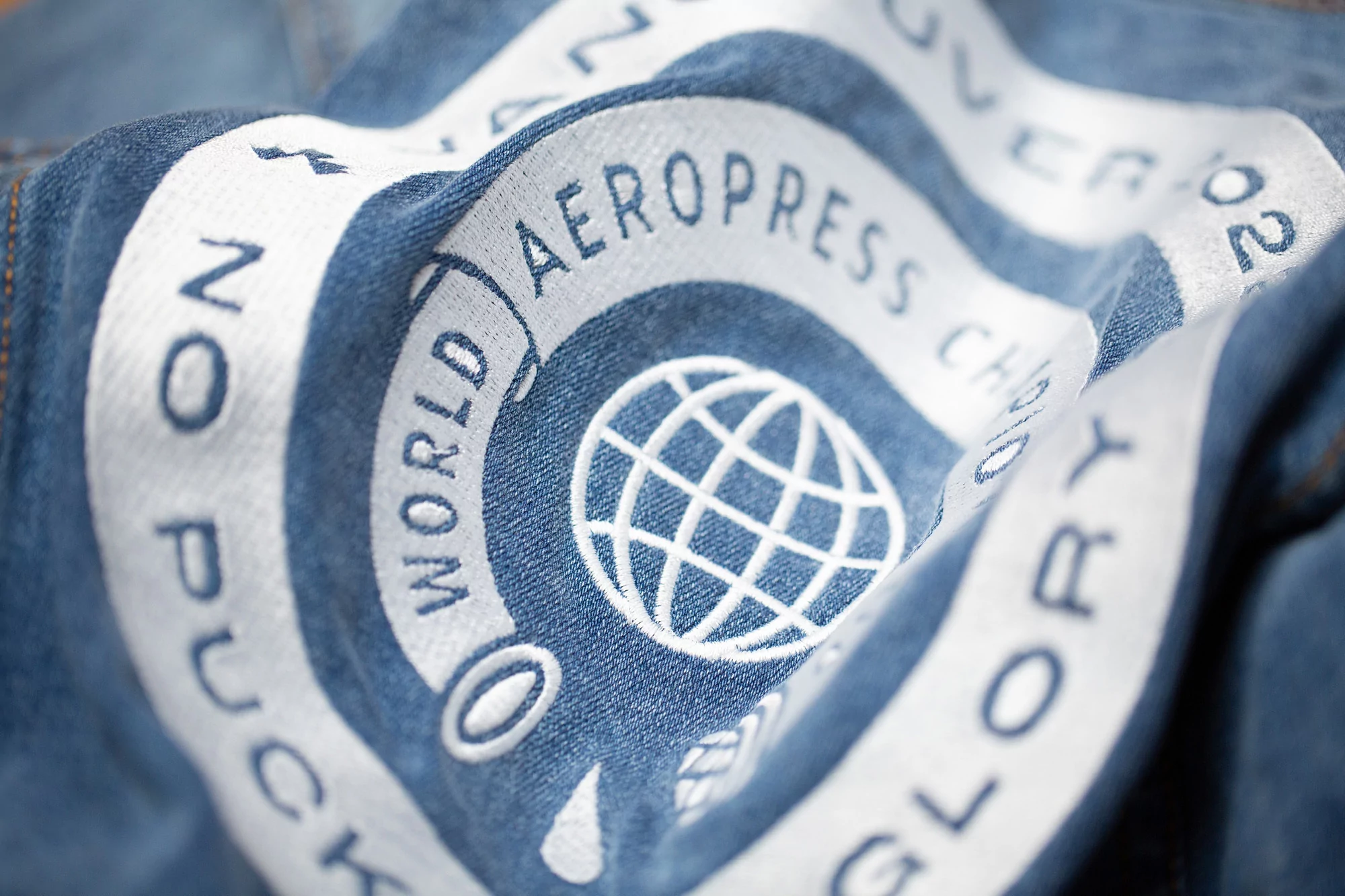 Close-up of blue fabric with white embroidered text and a globe design, possibly a coffee-related item.