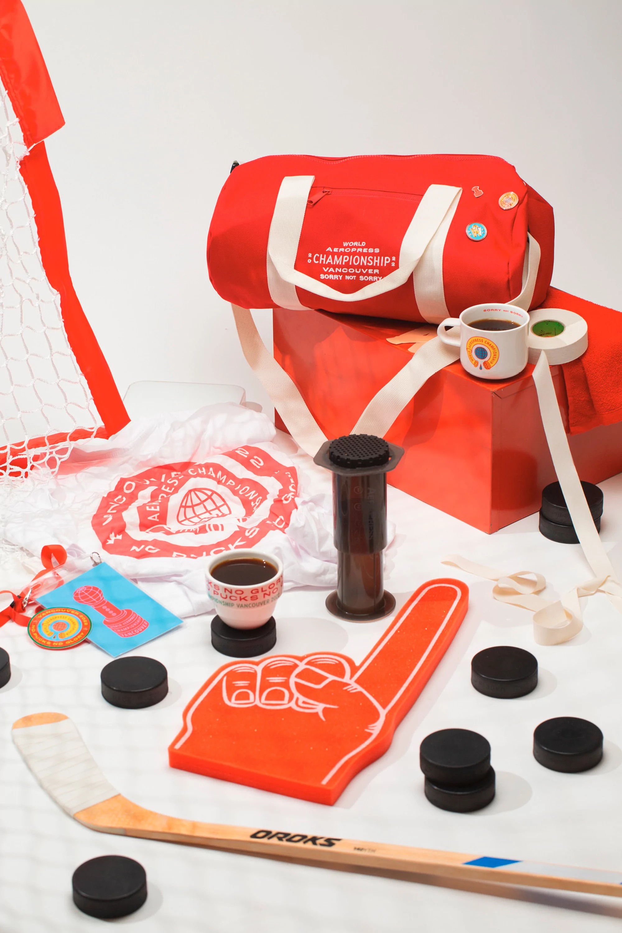 Still life featuring a red duffel bag, hockey equipment like a stick and pucks, and an Aeropress coffee maker against a white background.
