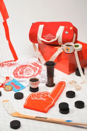 Still life featuring a red duffel bag, hockey equipment like a stick and pucks, and an Aeropress coffee maker against a white background.