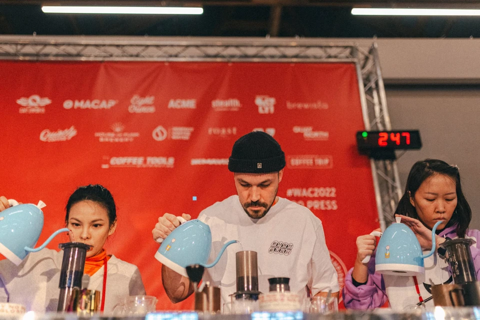 Three individuals are preparing coffee at a competition, using Aeropress equipment, in front of a branded red backdrop with digital timer.