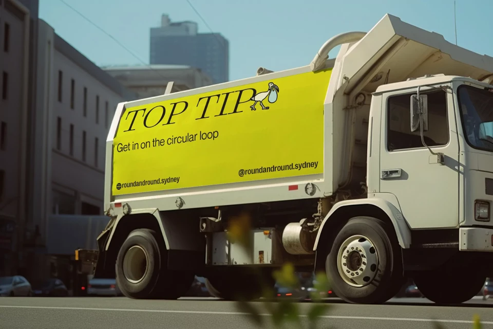 Garbage truck with yellow ad driving on city street. Signage reads 'Top Tip' and promotes sustainable waste management practices.