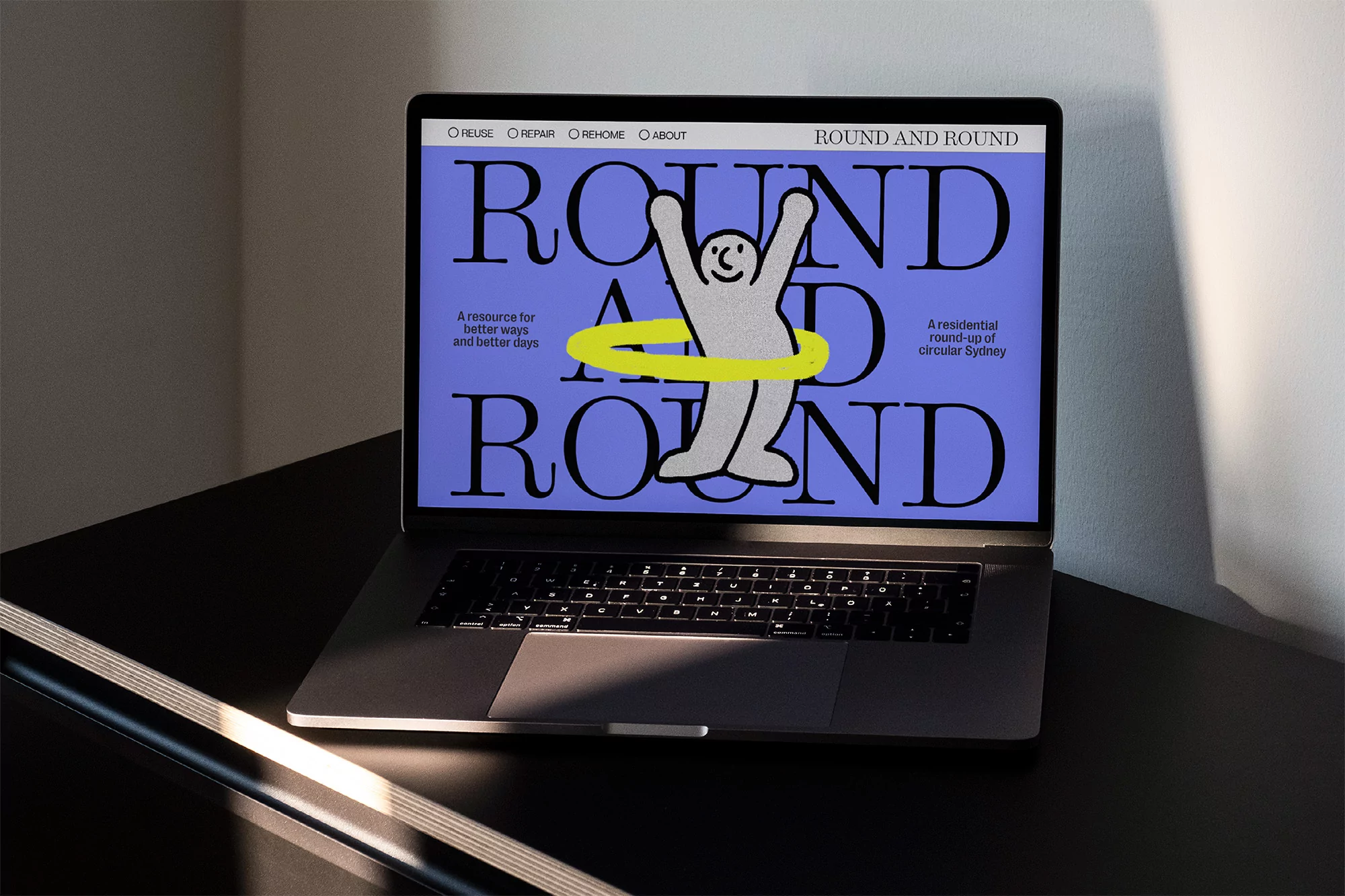 Laptop displays a website design with a figure inside a hoop, set on a dark desk against a plain background, illuminated by natural light.