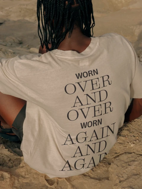Person from the back wearing a cream t-shirt with a dark inscription 'Worn over and over, worn again and again' sitting on sand.
