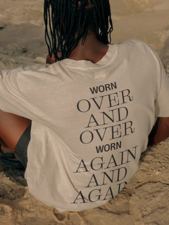Person from the back wearing a cream t-shirt with a dark inscription 'Worn over and over, worn again and again' sitting on sand.