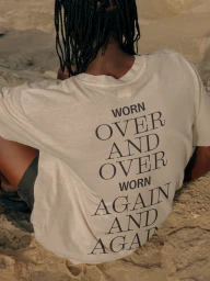 Person with braided black hair wearing a cream t-shirt with typography on the back, seated outdoors on sandy terrain.