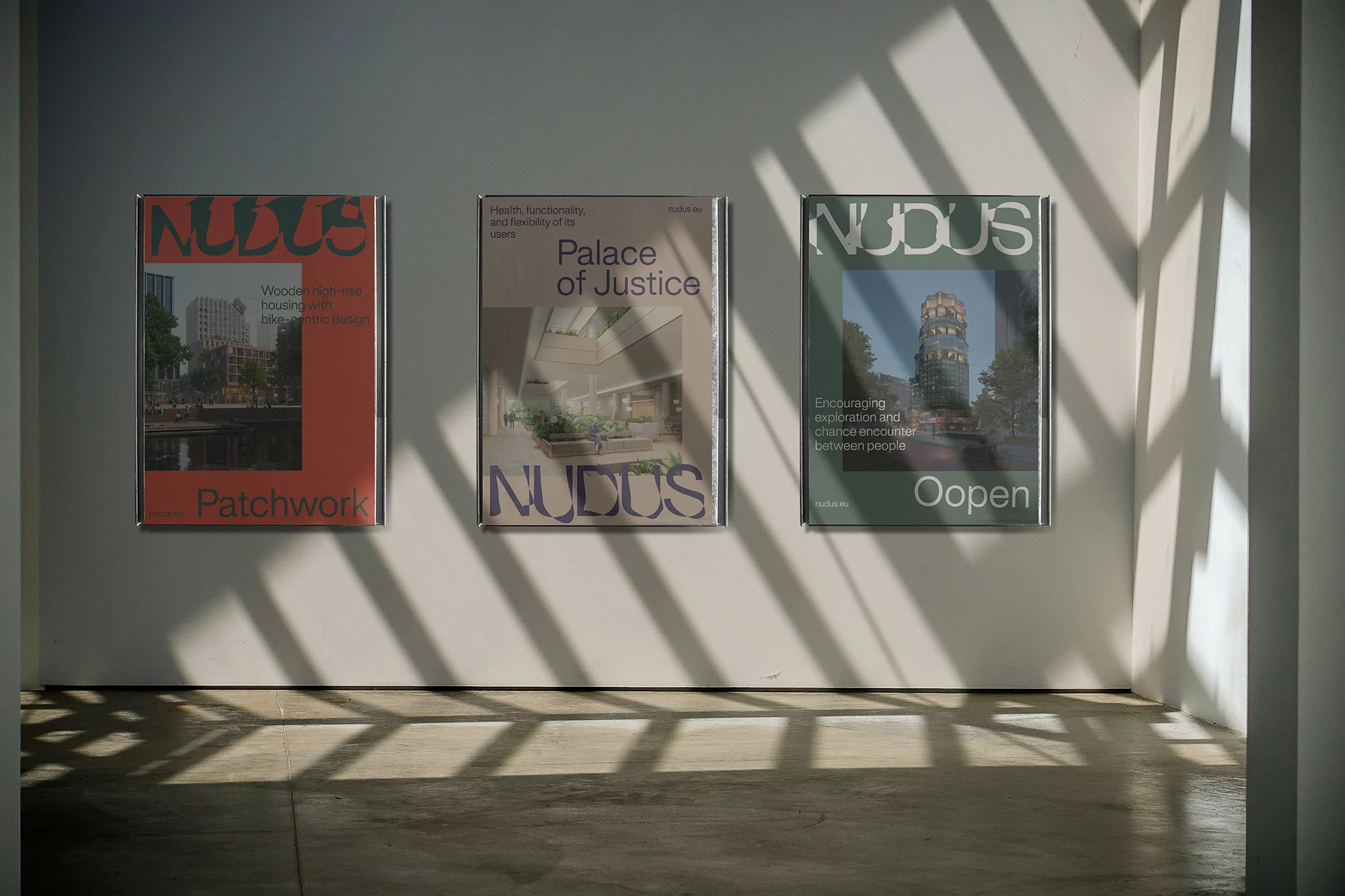 Three NUDUS architecture posters displayed on a white wall, catching bright sunlight, creating an indoor art gallery aesthetic.
