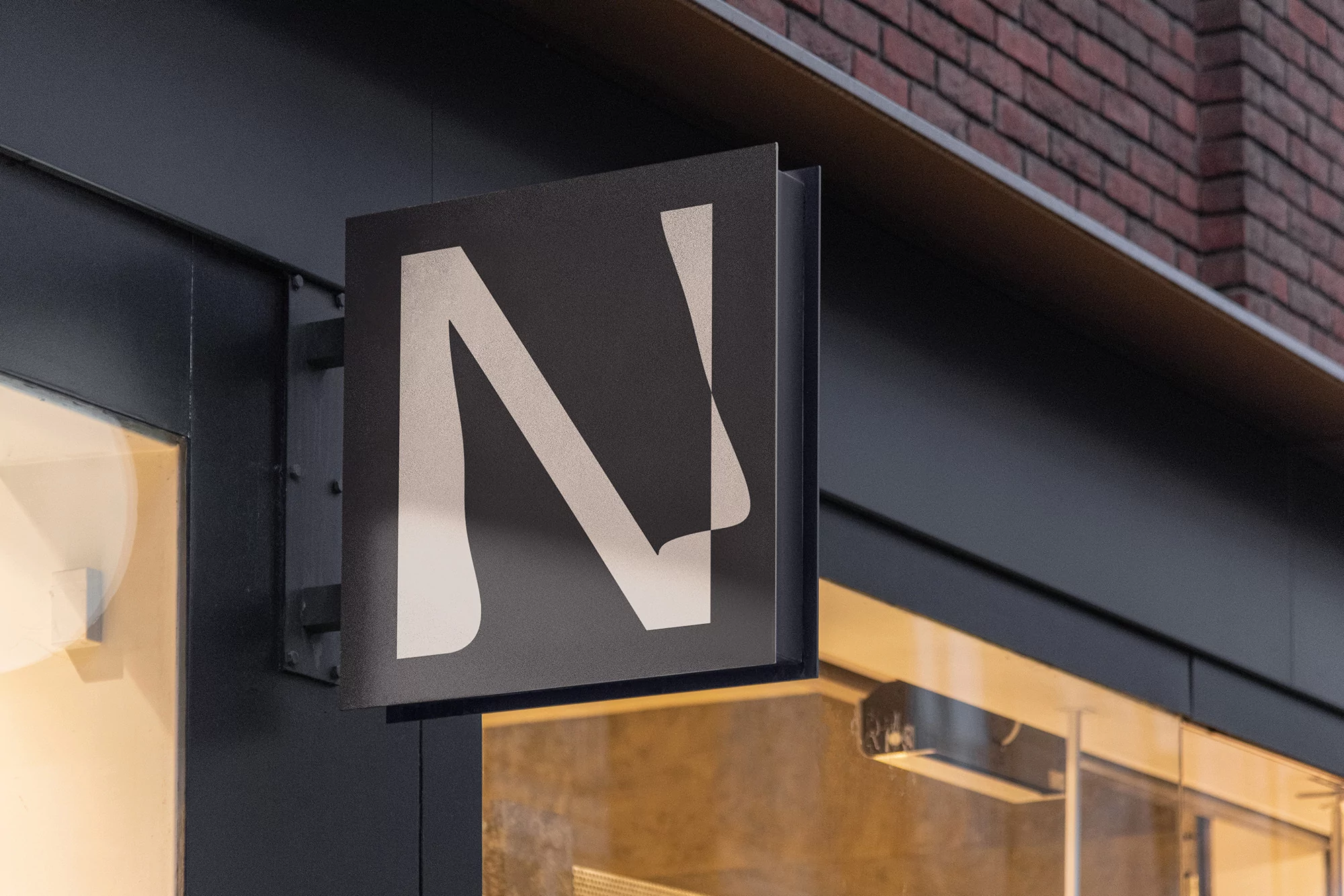 Illuminated business signage displays a simple, stylized letter N in light beige against a dark grey, mounted on a building exterior.