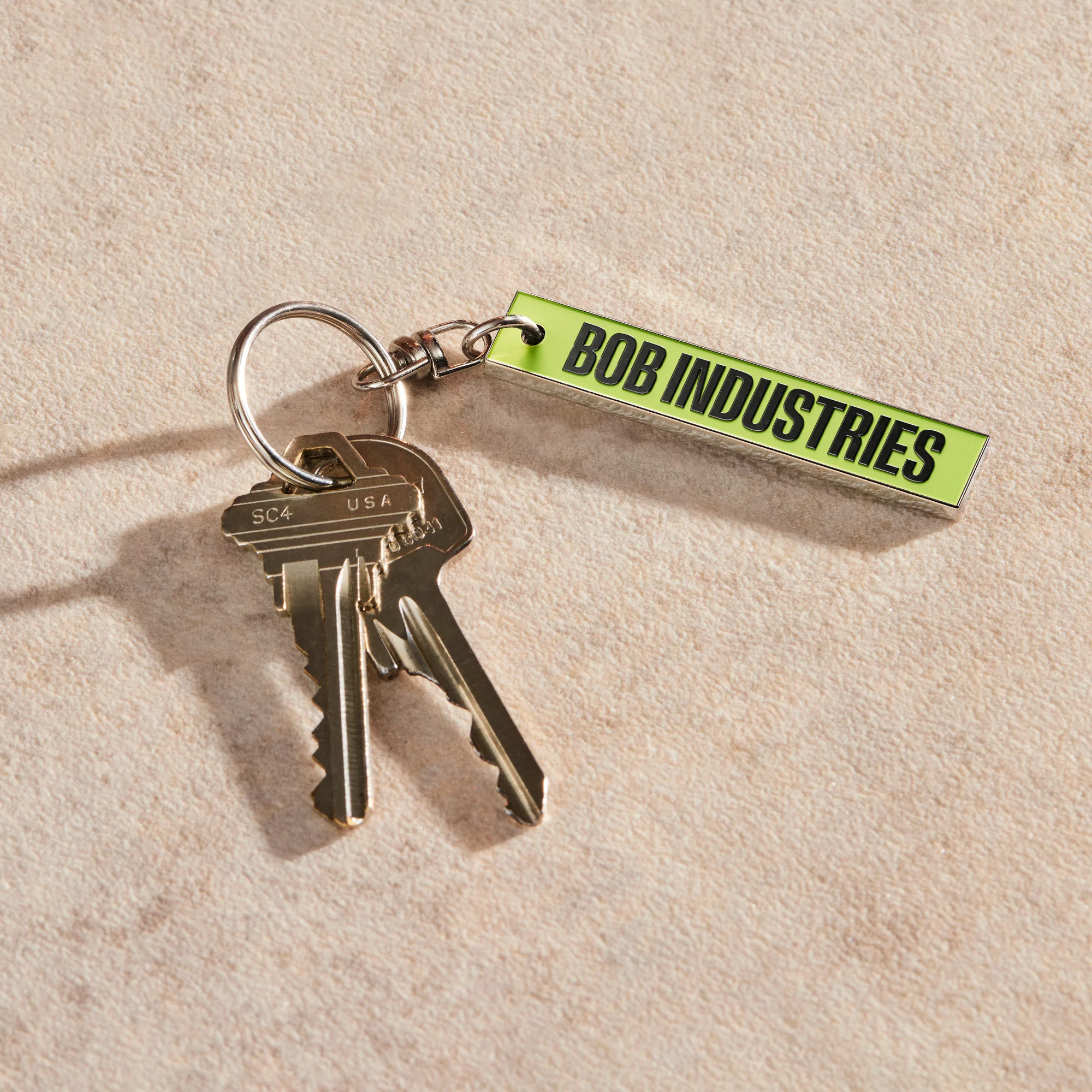 Close-up of keys on a ring with a neon green keychain reading 'BOB INDUSTRIES' against a neutral textured surface in a high-angle shot.