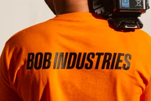Person wearing an orange t-shirt with black text Bob Industries on the back against a white wall.
