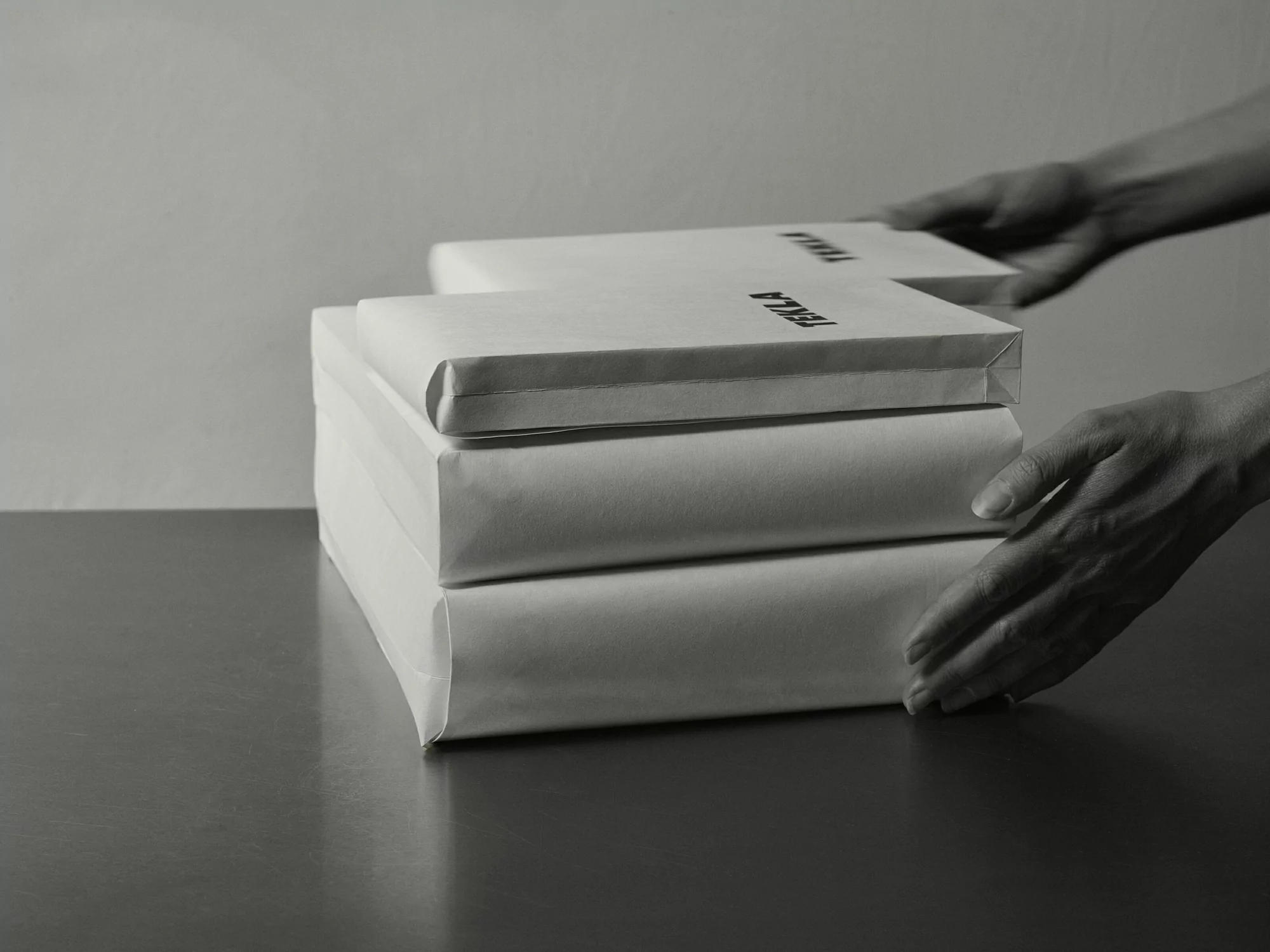 Stack of wrapped packages being held by fair-skinned hands in a black and white studio setting with soft, diffused lighting.