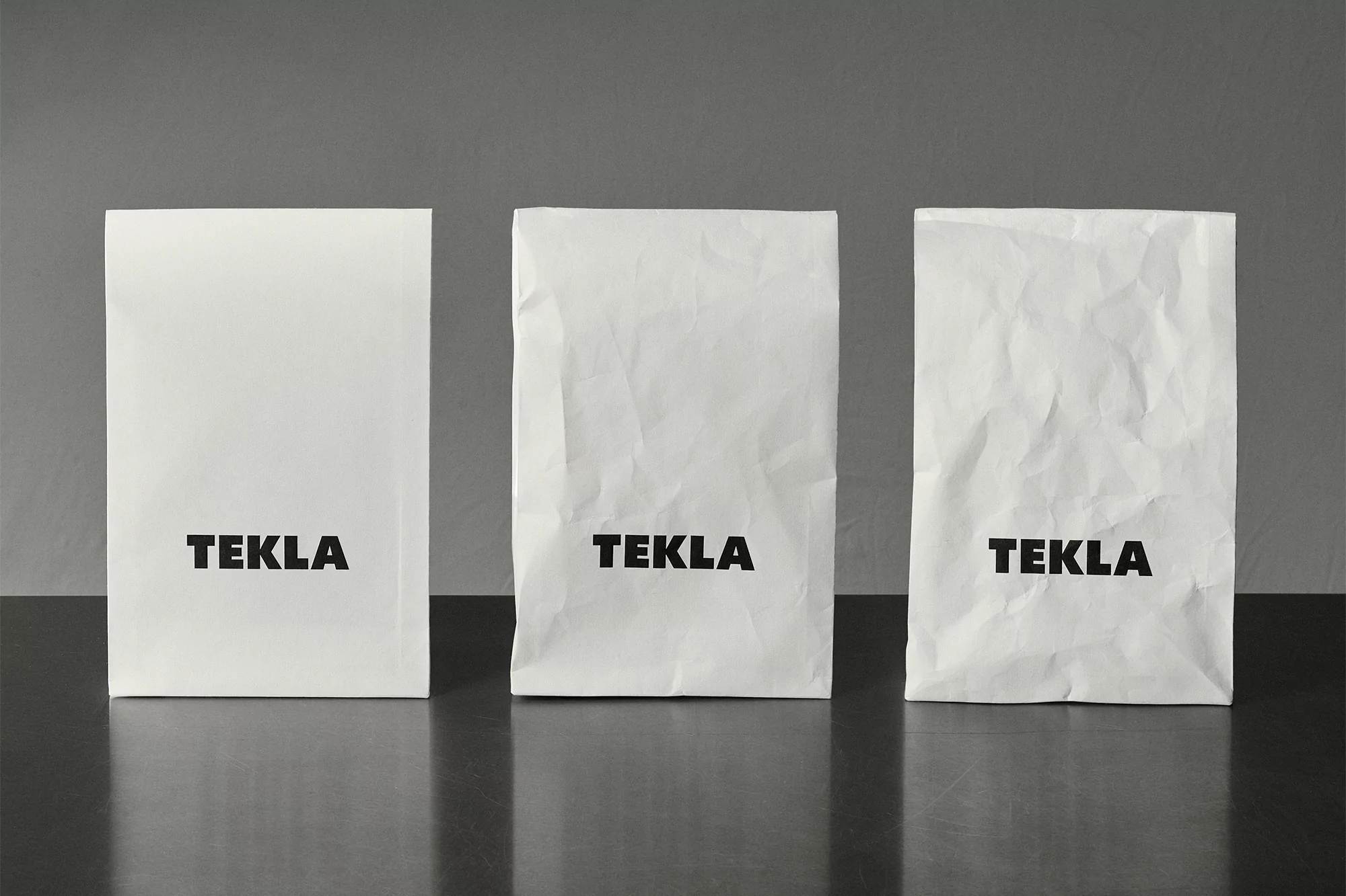 Three white paper bags with the word TEKLA in black font on a dark surface against a gray background in a studio shot.