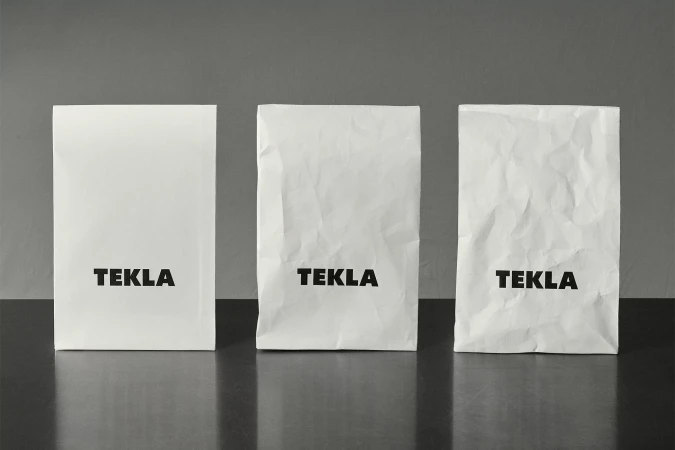 Three white paper bags with the word TEKLA in black font on a dark surface against a gray background in a studio shot.