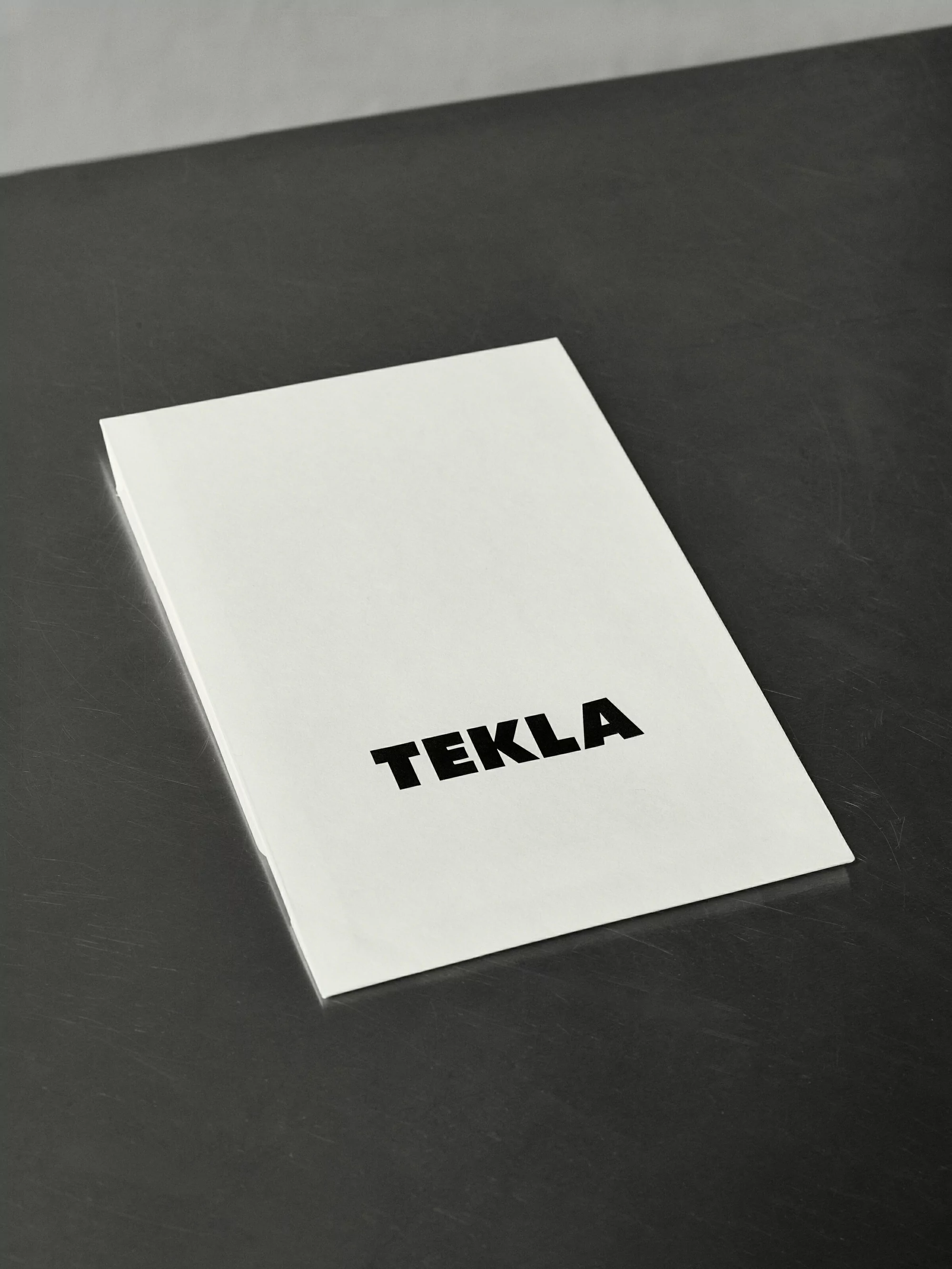 Minimalist photograph of a white TEKLA-branded booklet on a gray surface, ideal for showcasing product design and brand presentation.