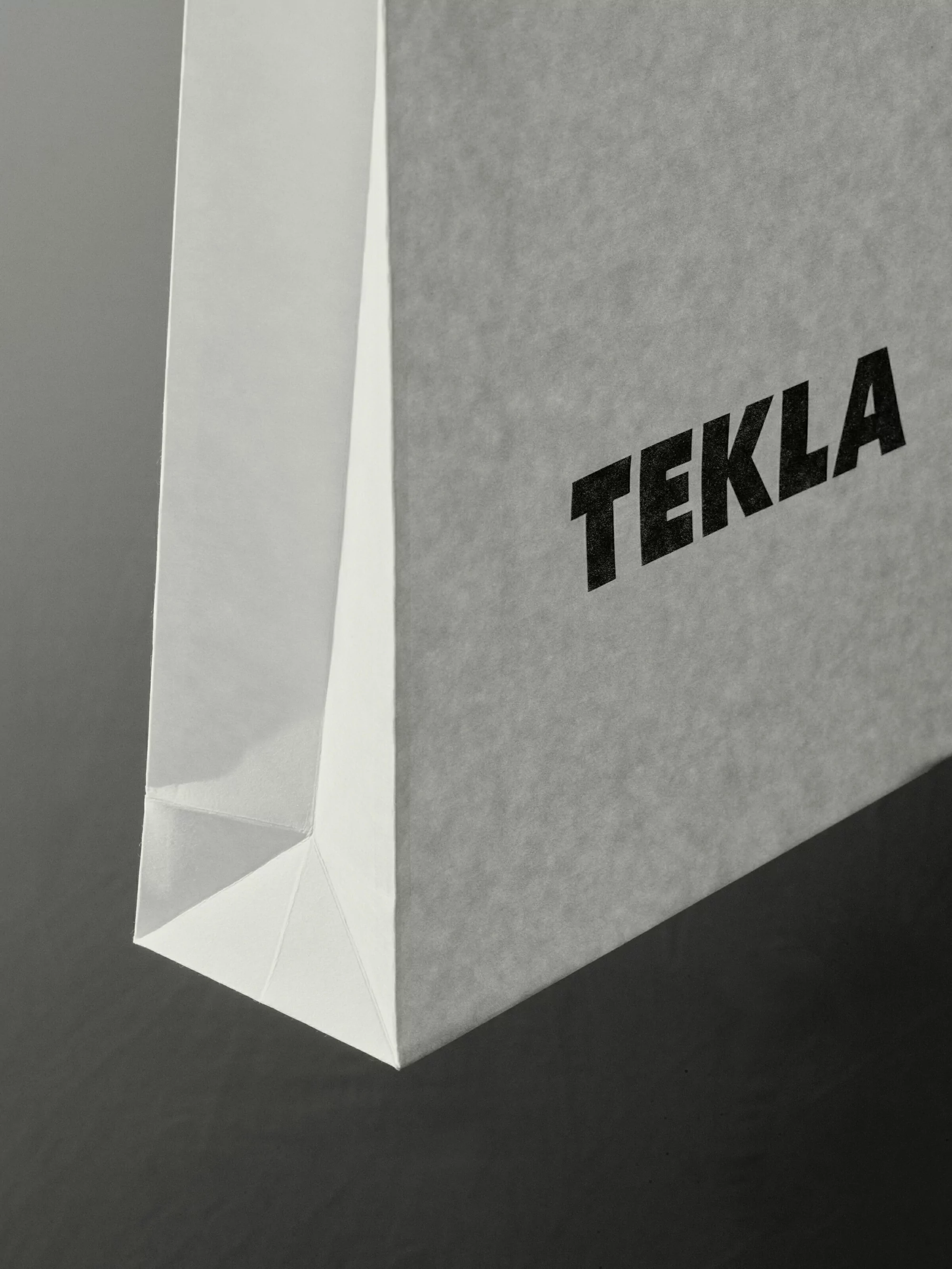 Close-up on a white paper Tekla shopping bag with a clean, minimalist design against a dark gray background, accentuating its texture.