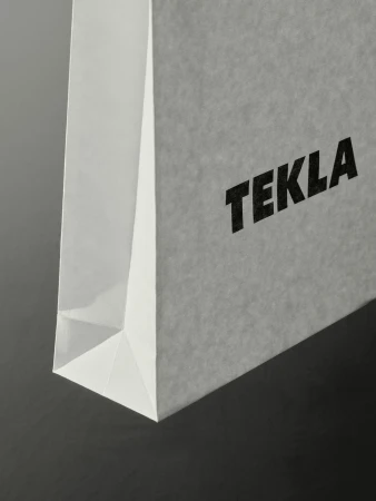 Close-up on a white paper Tekla shopping bag with a clean, minimalist design against a dark gray background, accentuating its texture.