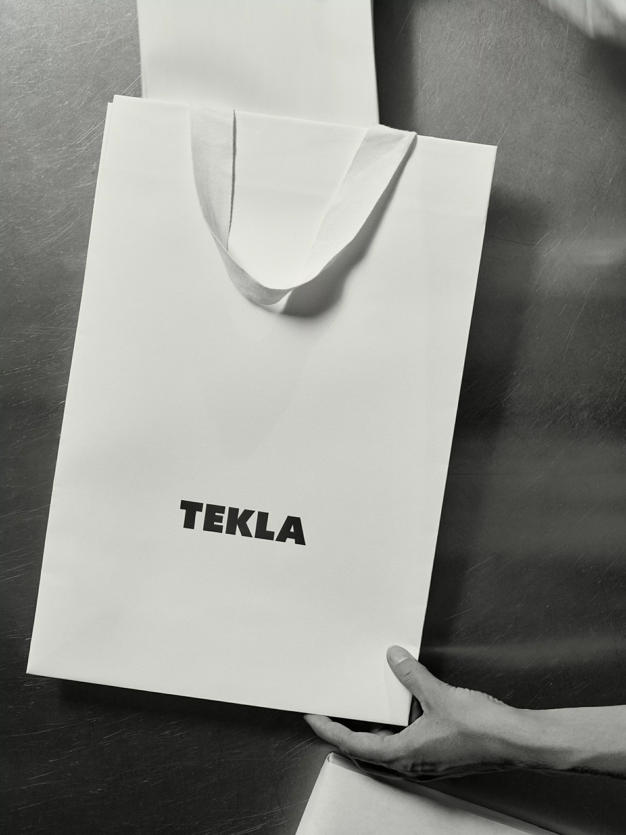 A white shopping bag with black 'TEKLA' lettering, held by a hand against a grey background, suggesting retail or branding context.