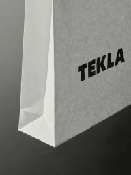 Textured white paper bag with bold black Tekla branding resting on a dark surface.