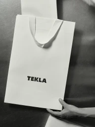 White paper shopping bag with Tekla branding held against a dark textured background