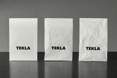Three white paper bags with bold black TEKLA branding standing upright on a dark reflective surface against a grey backdrop