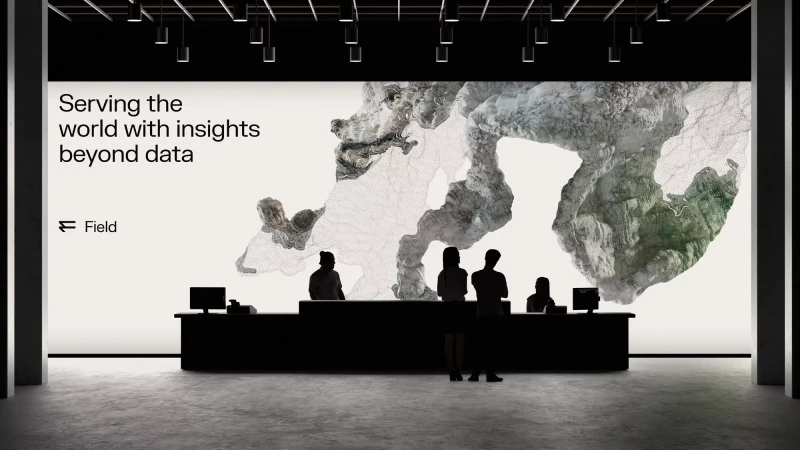 People at a dark reception desk with computer monitors, silhouetted against a backdrop featuring abstract cloud-like art and text.