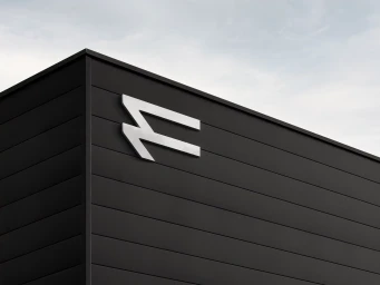White metallic brand logo mounted on a dark horizontal panel exterior wall against an overcast sky