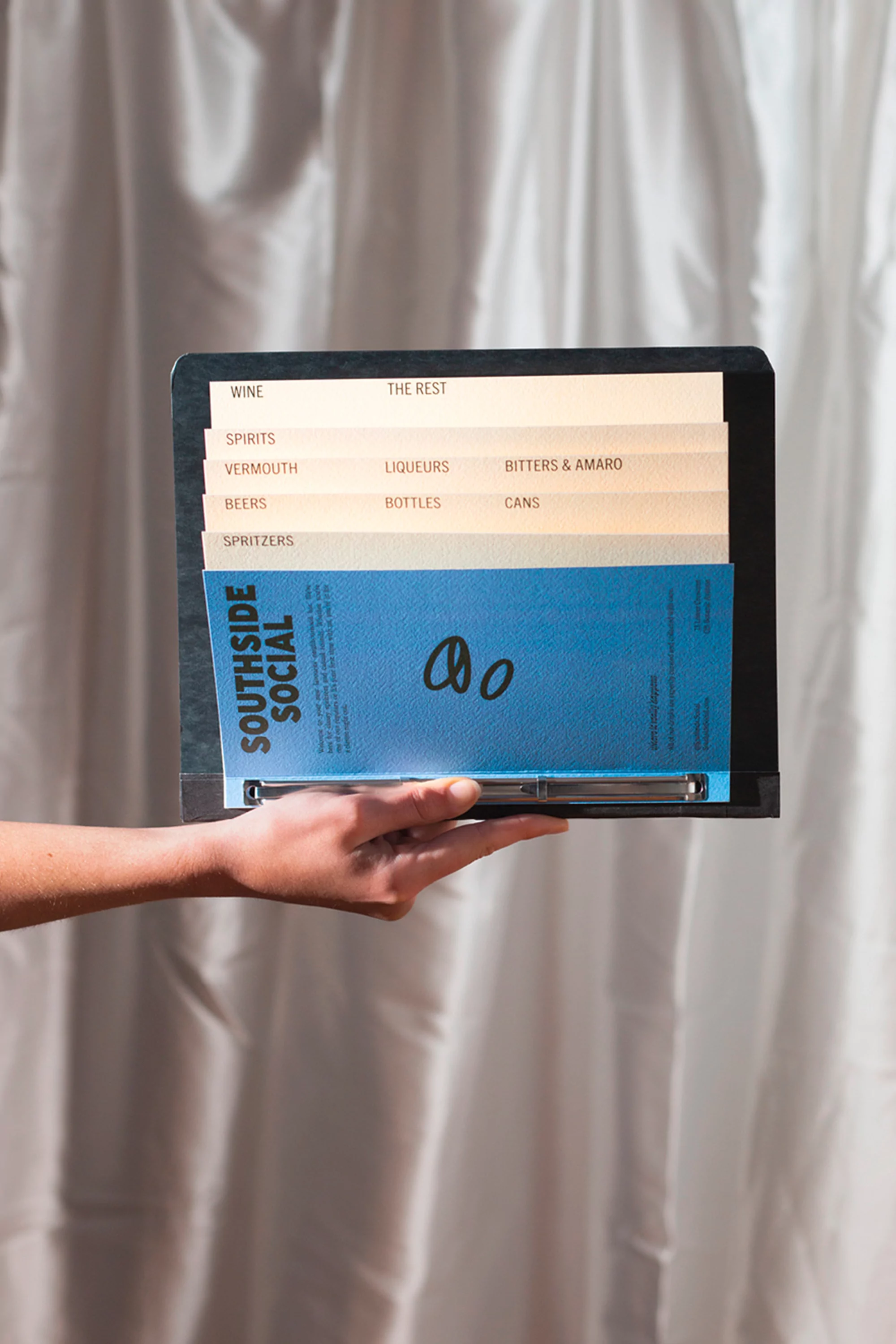 Hand holding black menu holder with blue Southside Social menu insert and menu dividers against white draped fabric backdrop.