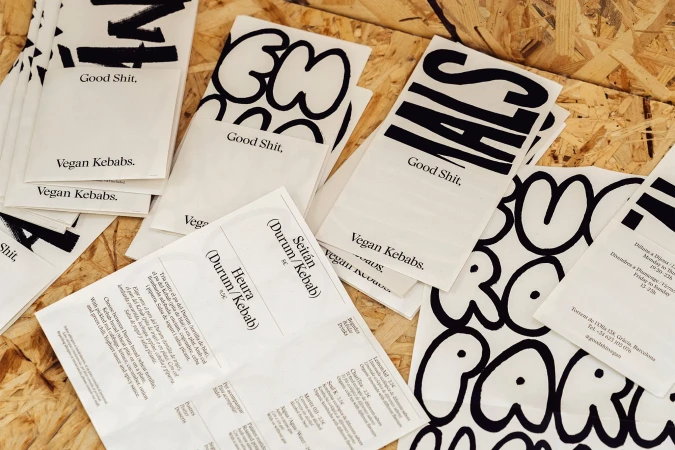 Promotional materials for vegan kebabs on a wooden surface, featuring bold black and white graphics and menu details.