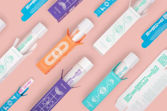 Top-down view of colourful cosmetic product packaging and bottles featuring geometric graphic branding on a pink background.