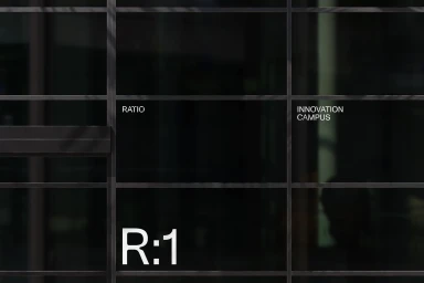 Dark glass facade with a dark metal grid frame displaying white typography reading RATIO, INNOVATION CAMPUS, and R:1.