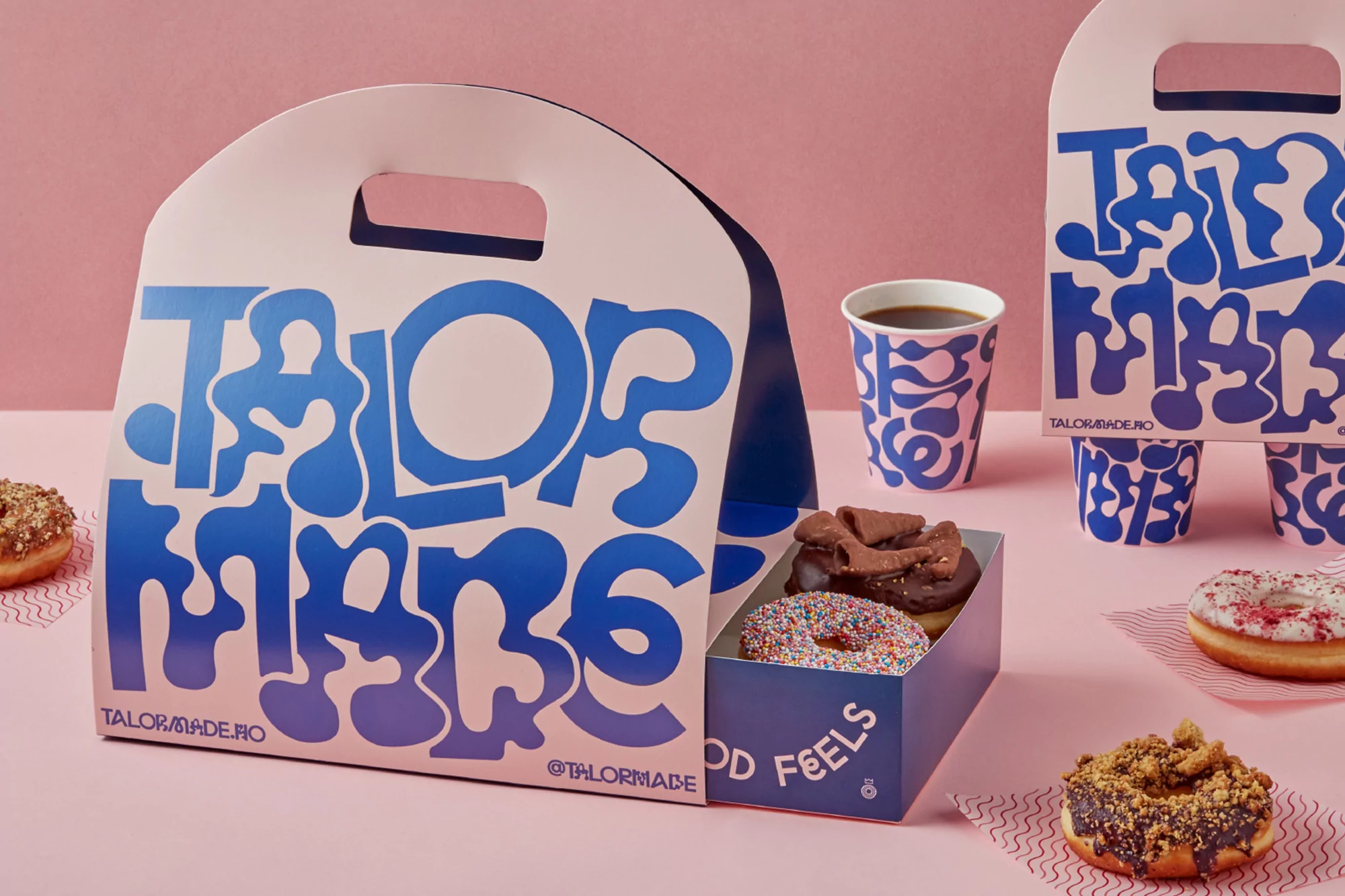Donuts in a branded box and cup with the logo 'Tailor Made' on a pink background, alongside other donuts on patterned papers.
