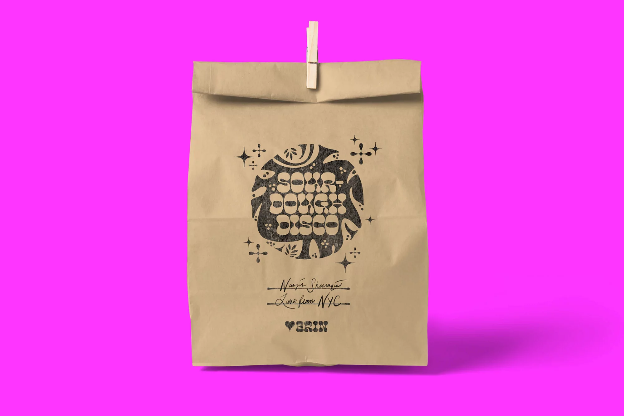Paper bag with a black graphic design and text on a bright pink background, held by a wooden clip.