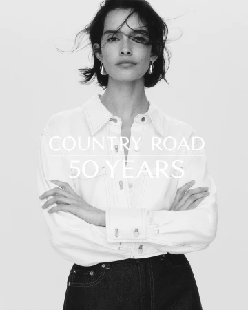 Monochrome portrait of a brunette woman in a white shirt and jeans with silver earrings, posing with crossed arms indoors.