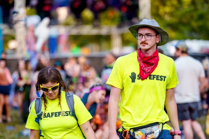 Two people at an outdoor event, wearing bright yellow SAFEFEST t-shirts, with a blurred crowd in the background on a sunny day.